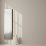 Halbtransparente Gardine vor einem Fenster diffundiert Tageslicht, ein schlanker Wandspiegel und eine weiße Keramikvase verstärken die Lichtlenkung.