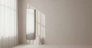 Halbtransparente Gardine vor einem Fenster diffundiert Tageslicht, ein schlanker Wandspiegel und eine weiße Keramikvase verstärken die Lichtlenkung.